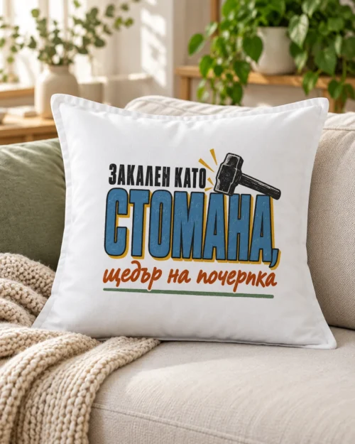 Бяла възглавница с едри букви и надпис Закален като стомана, щедър на почерпка – на дивана