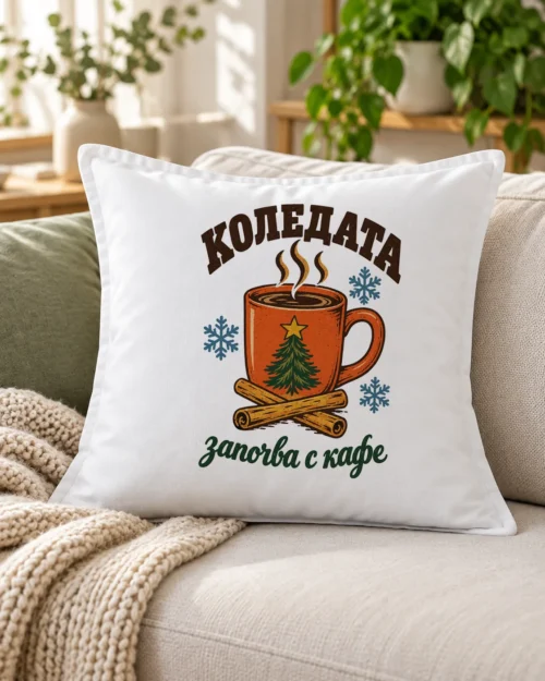 Бяла възглавница с чаша кафе, елхова клонка, канела и надпис Коледата започва с кафе – на дивана