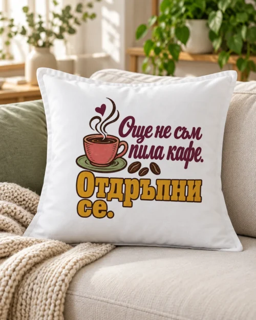 Бяла възглавница с чаша кафе, зърна, сърце и надпис Още не съм пила кафе – на дивана